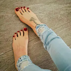Barfuss mit roten Zehennägeln und ihrem Fuss-Tattoo in zerrissenen Jeans auf grauem Boden - Fussfetisch Zürich